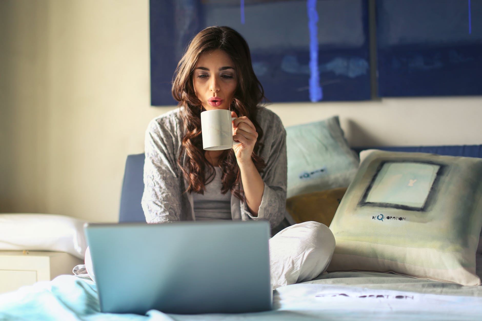 woman in grey jacket sits on bed uses grey laptop